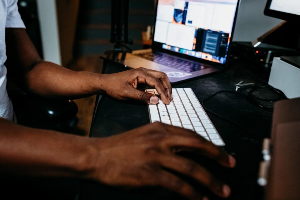 Ein Mann schreibt etwas auf seiner Tastatur, im Hintergrund ist ein Computerbildschirm zu sehen und der Raum ist dunkel.